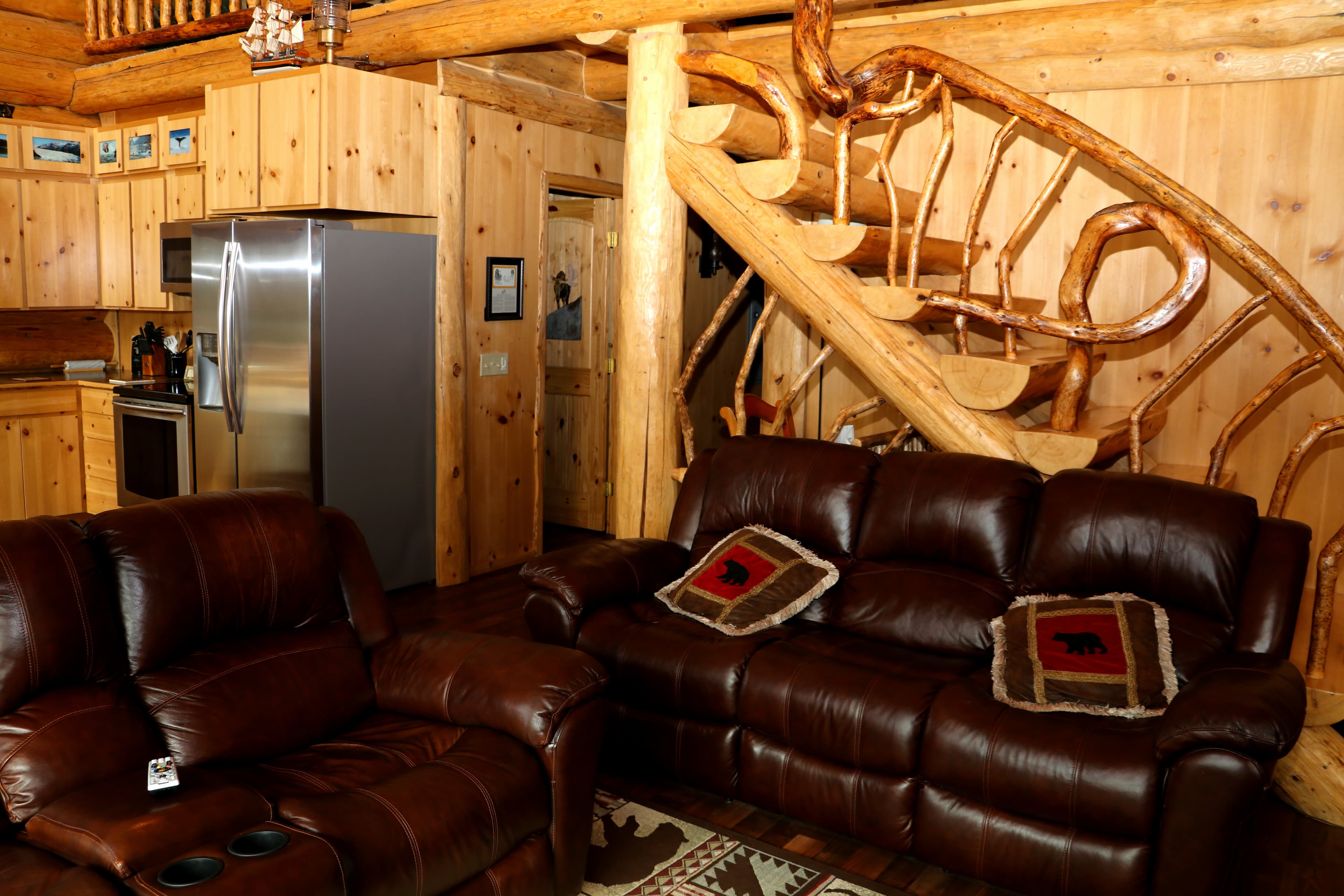First Floor View 2 - sitting area with rich brown leather couch and loveseat near bottom of stairs with rustic handrailing, kitchen is visible in background.