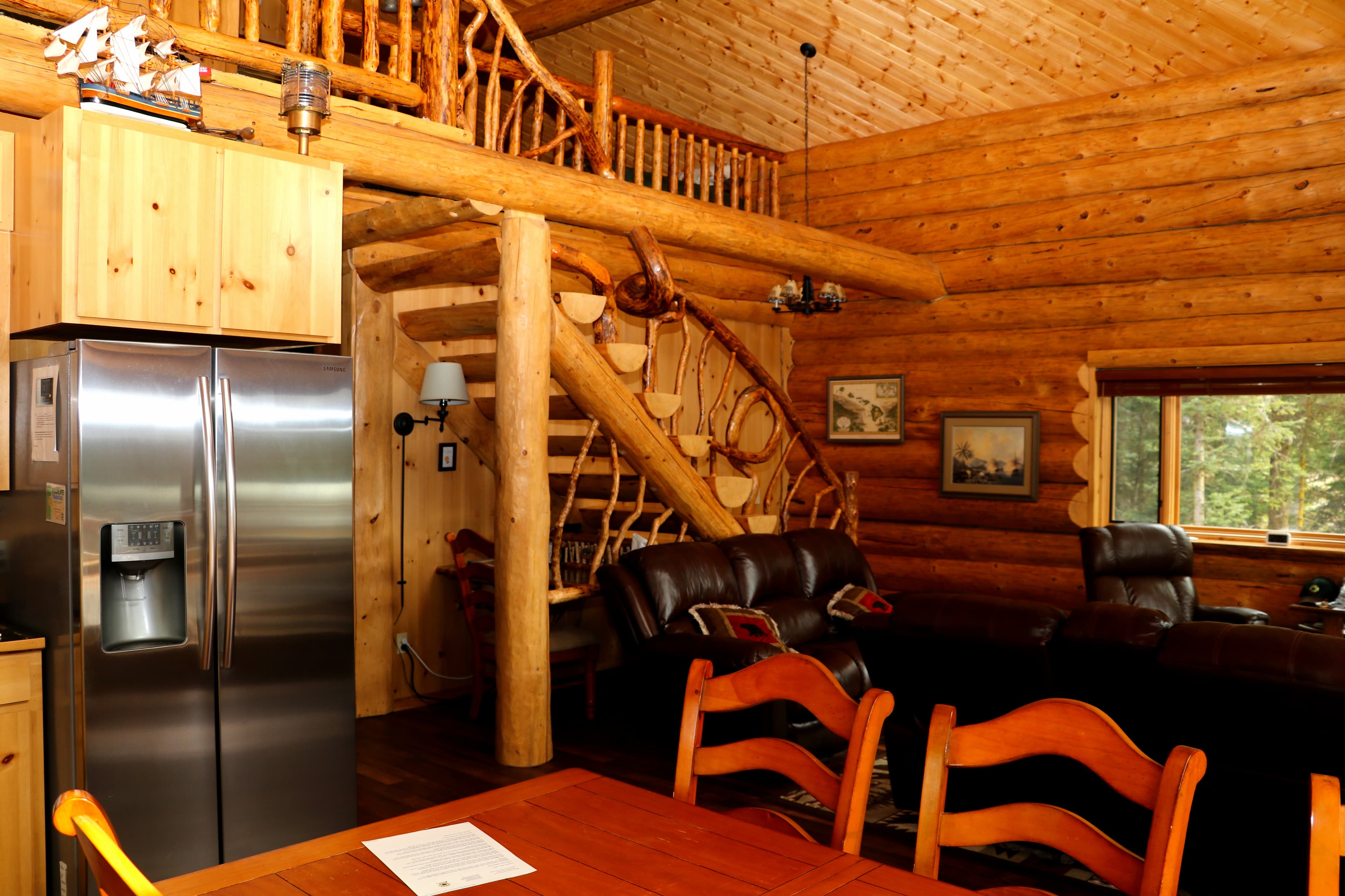 First Floor View 4 - dining area in foreground with stainless steel refrigerator in open kitchen and sitting area in background with stairs leading to loft.