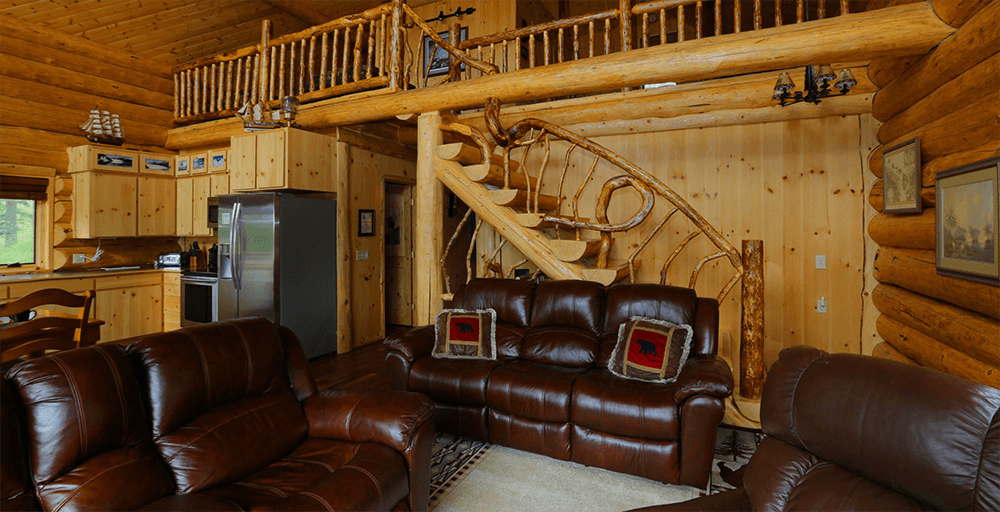 A living room inside of a log cabin featuring reddish brown leather sofa, loveseat, and chair with an intricately carved staircase leading up to a loft