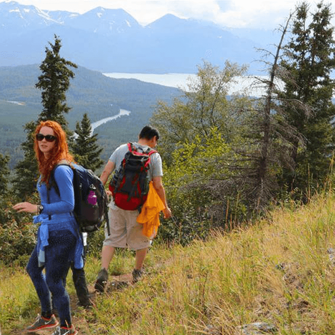 Two hikers navigating a trail with mountains and a river in the background.