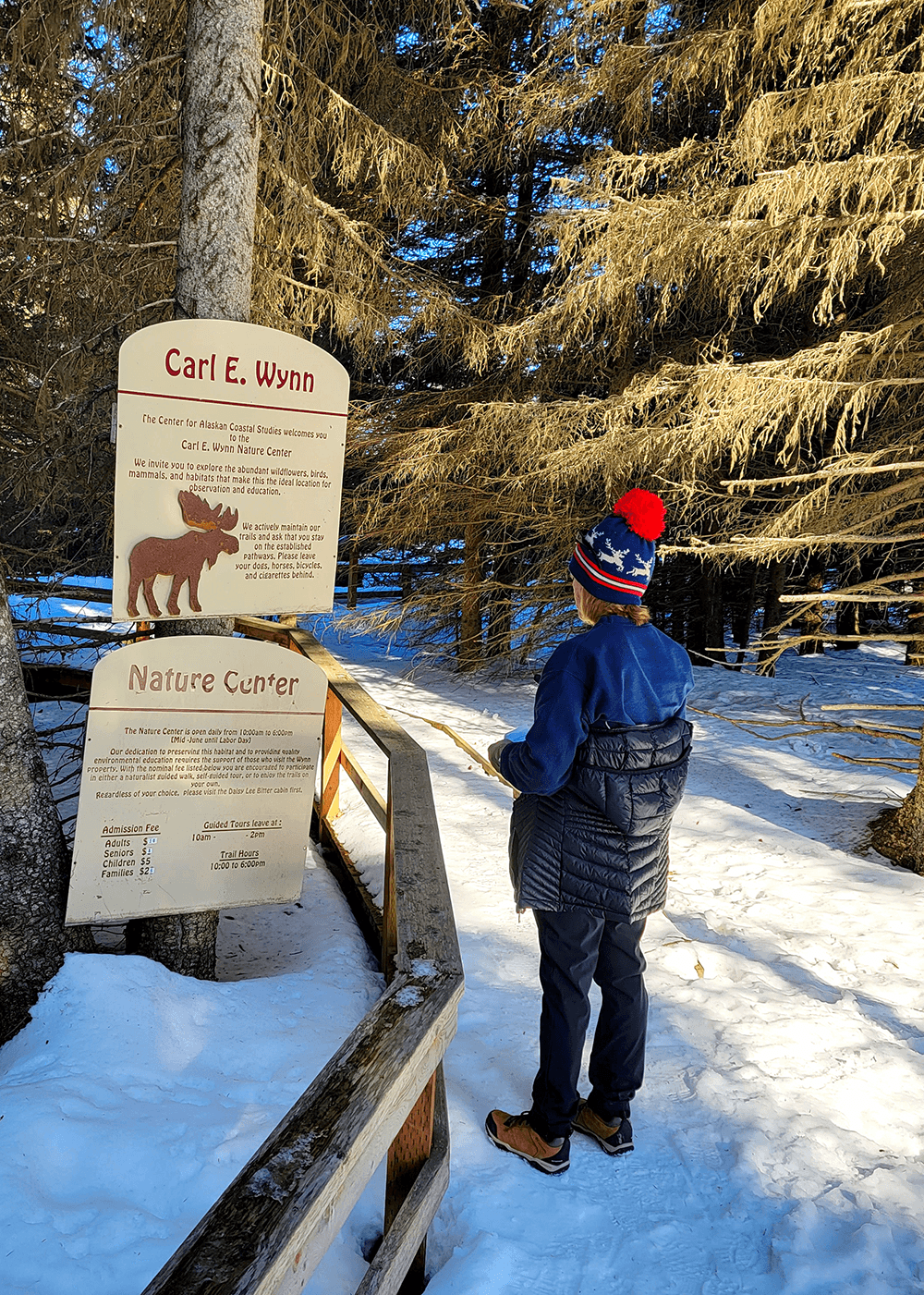 A person in a winter hat stands in front of a nature center sign surrounded by snow-covered trees.