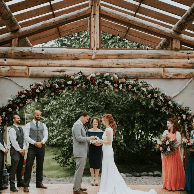 A bride and groom exchange vows under a floral arch during their wedding ceremony, surrounded by guests.