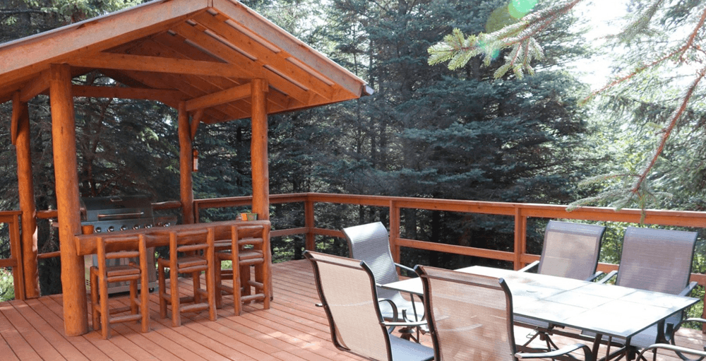A wooden deck with a grilling area and seating, surrounded by tall trees.