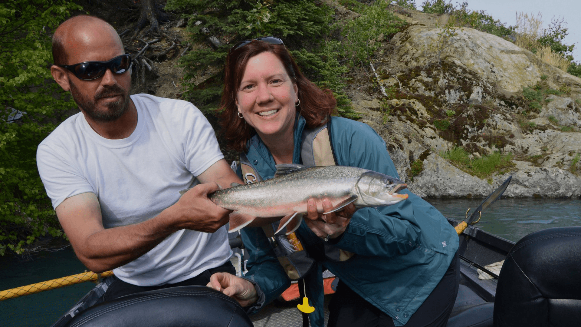 A man and woman smile while holding a fish on a boat surrounded by trees and rocks.