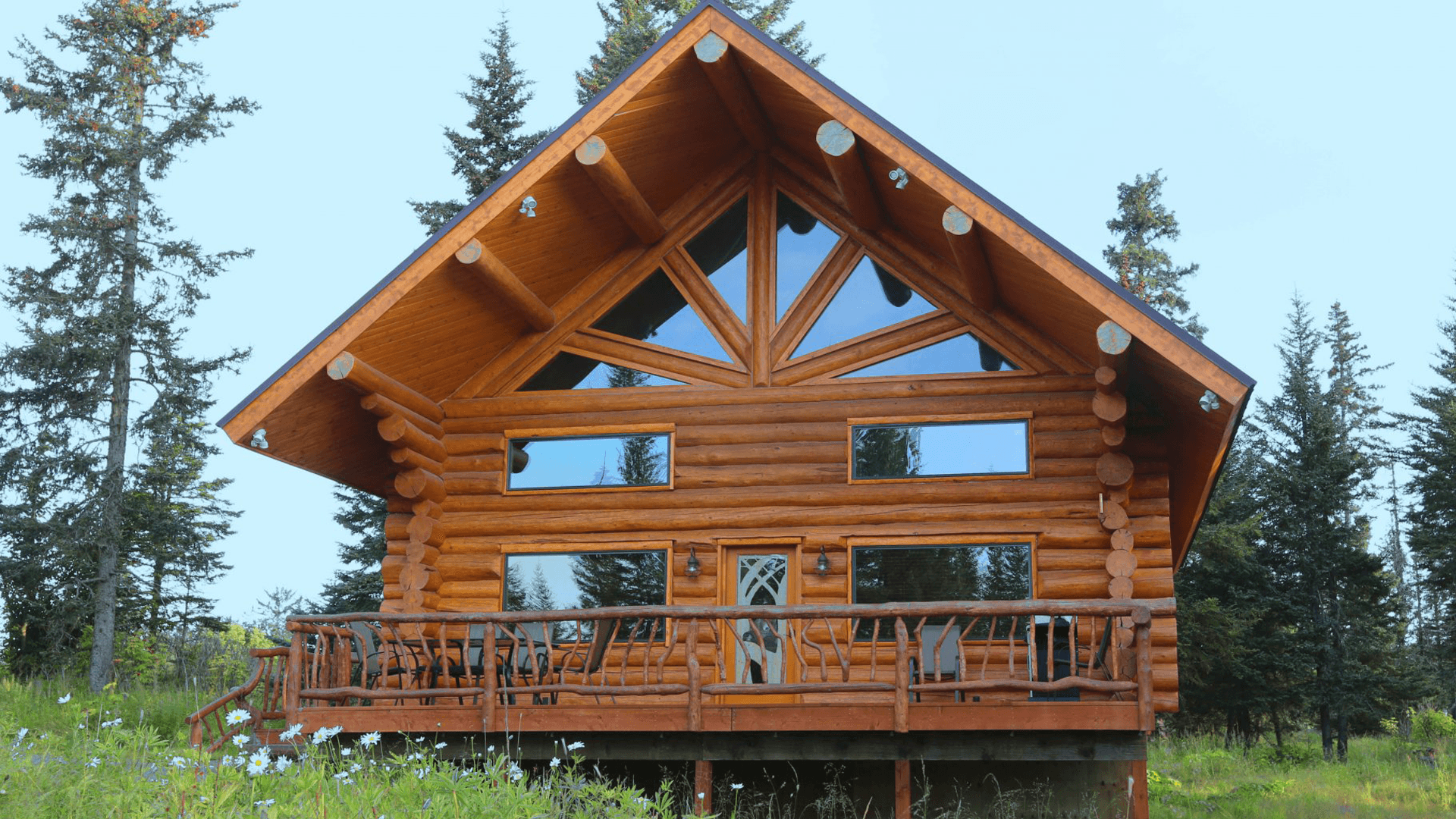 Exterior of a log cabin with a large, triangle shape of windows on a blue-sky day with green grass and wildflowers in the foreground.